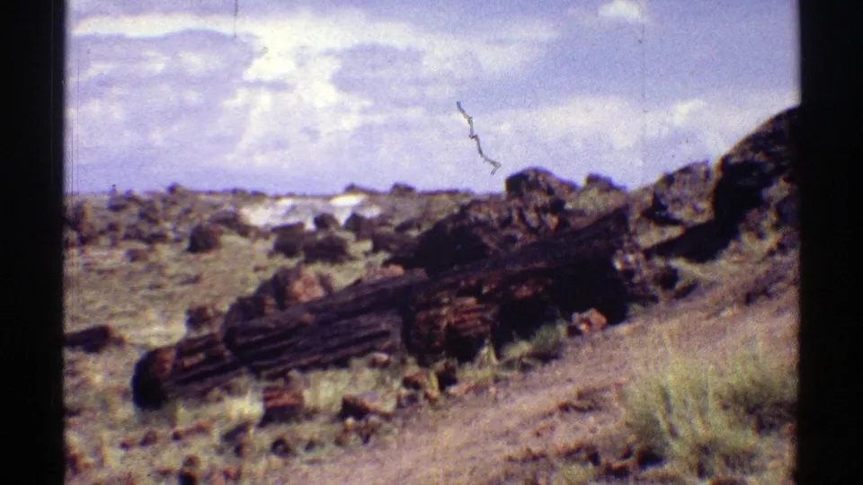 1969 Arizona home-movie frame showing dark rock in the foreground, scattered desert stones, dry grass, and a pale cloud-filled sky.