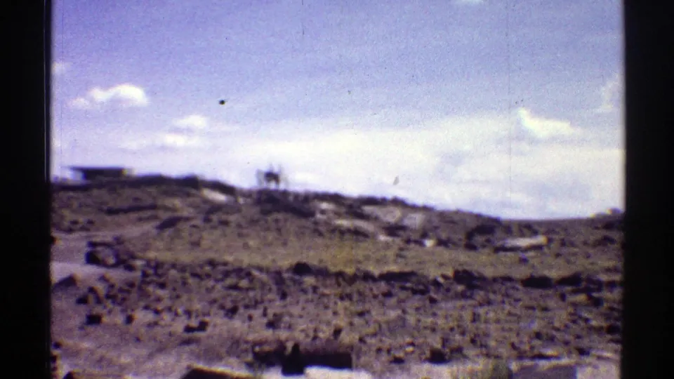 Wide 1969 Arizona home-movie view across rocky arid ground toward low hills and a small distant structure or vehicle under pale clouds.