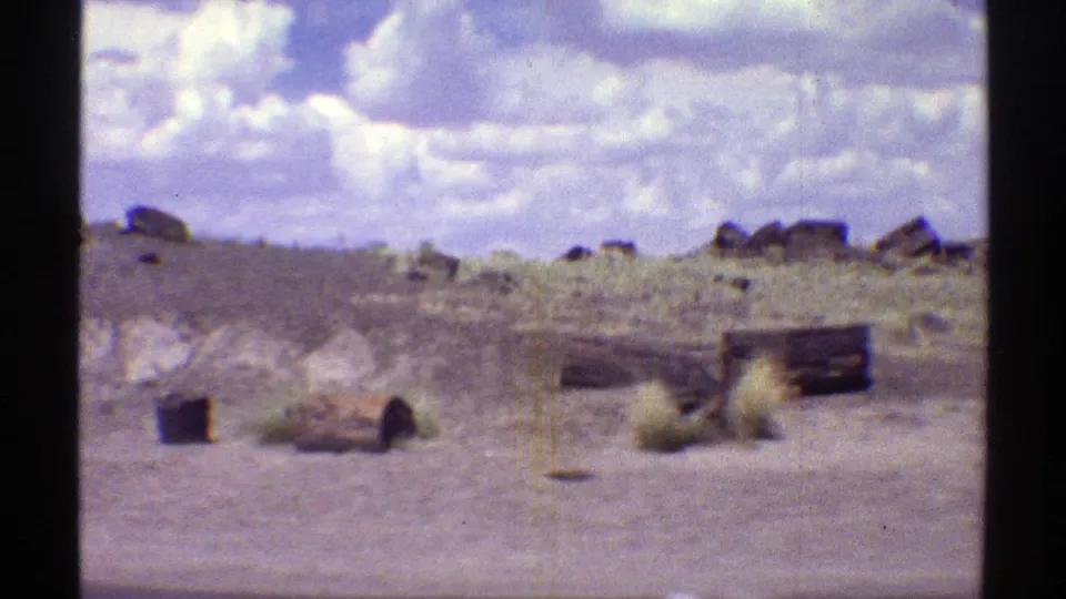 1969 Arizona desert roadside view with pale rocky ground, scrub plants, low rocks, and a distant white and tan camper-like vehicle.