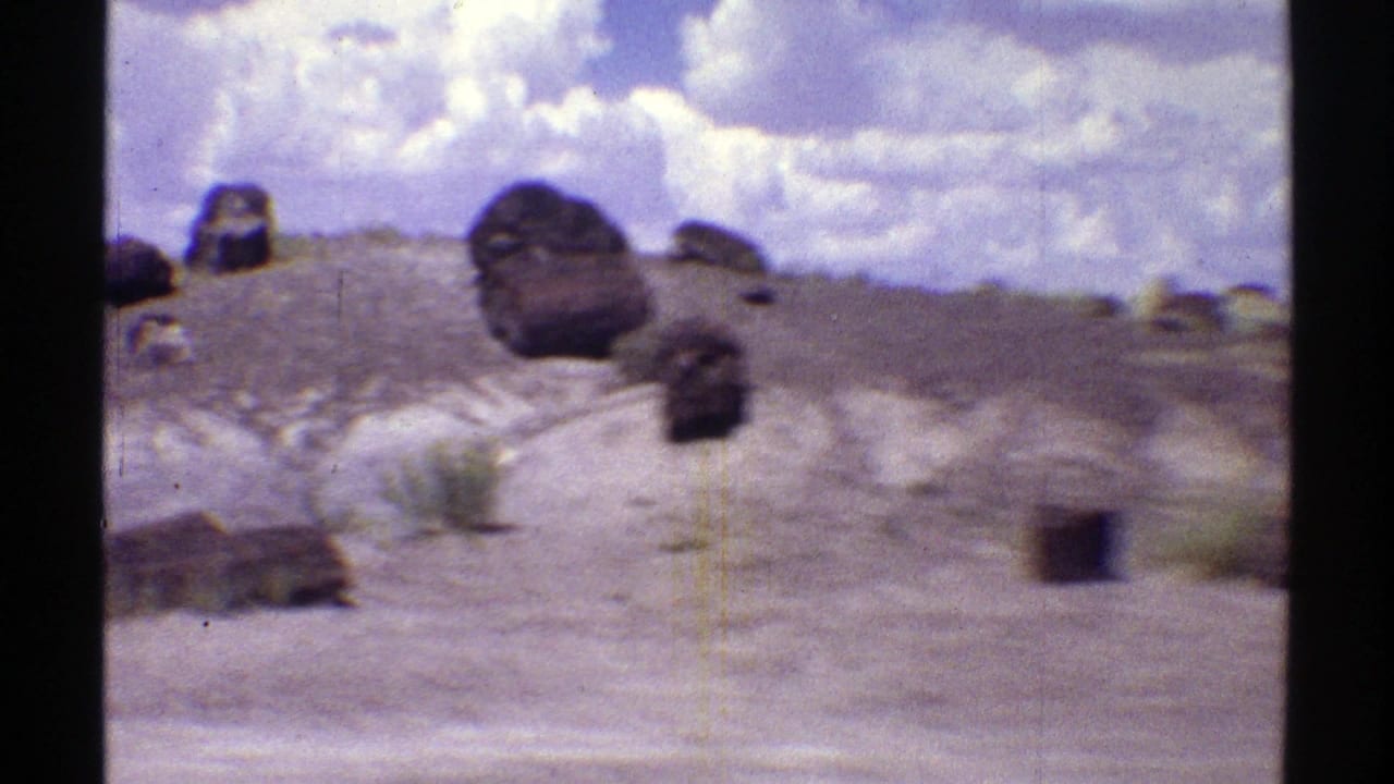 1969 Arizona 8mm frame showing pale desert terrain with scattered rounded stones, low shrubs, and clouds overhead.