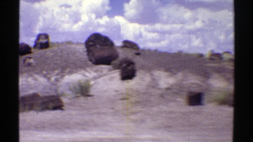 1969 Arizona 8mm frame showing pale desert terrain with scattered rounded stones, low shrubs, and clouds overhead.