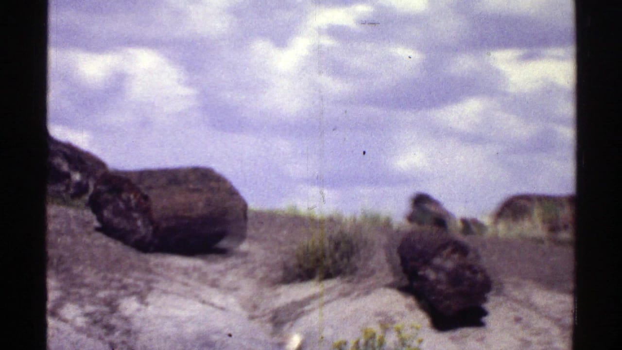 1969 Arizona desert view with dark boulders, scattered brush, pale soil, and a cloud-filled sky.