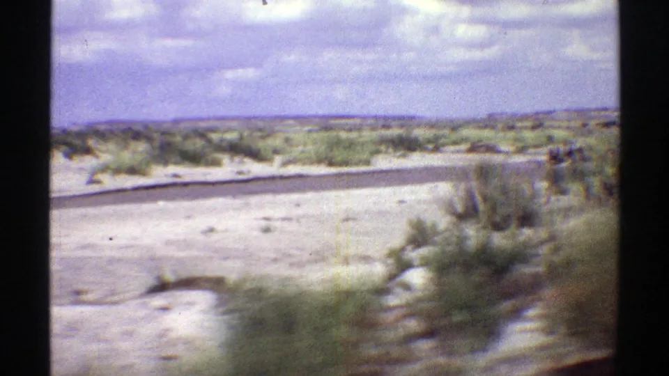 Wide 1969 Arizona desert frame showing pale open ground, scattered shrubs, distant low terrain, and cloudy sky.