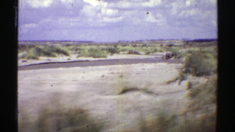 1969 Arizona 8mm roadside view from a moving vehicle, showing sandy terrain, low shrubs, and a paved road.