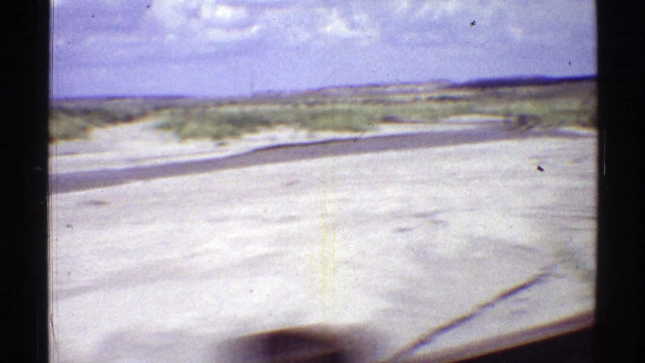 Side-window view of an Arizona desert road in 1969, with pale sand, scattered brush, and distant low hills.