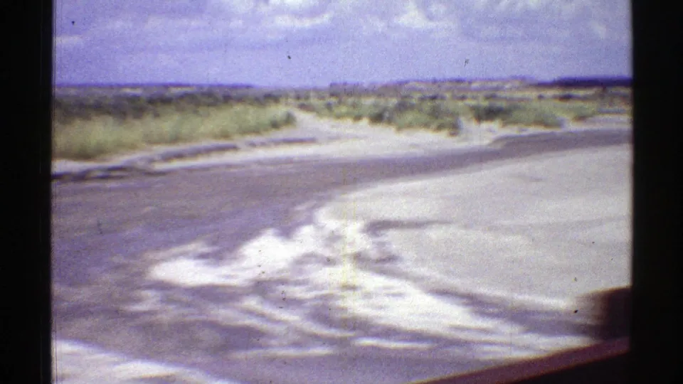 1969 Arizona roadside footage showing a curve or wash beside the pavement, with shrubs and open desert beyond.