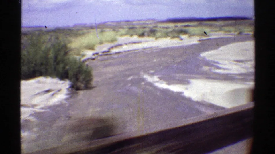 Passenger-side 8mm view in Arizona, 1969, with desert shrubs, sandy road edge, and the vehicle window frame visible.