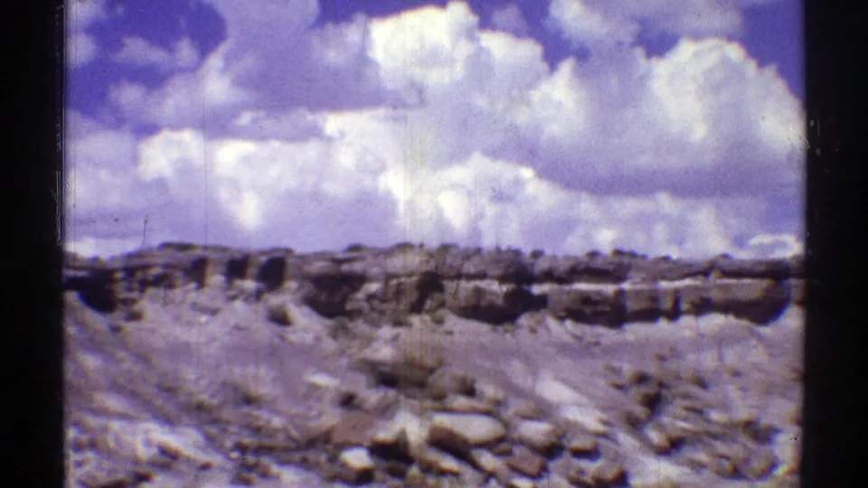 Arizona desert landscape with scattered rocks, low ridges, and large white clouds captured on soft 8mm film.