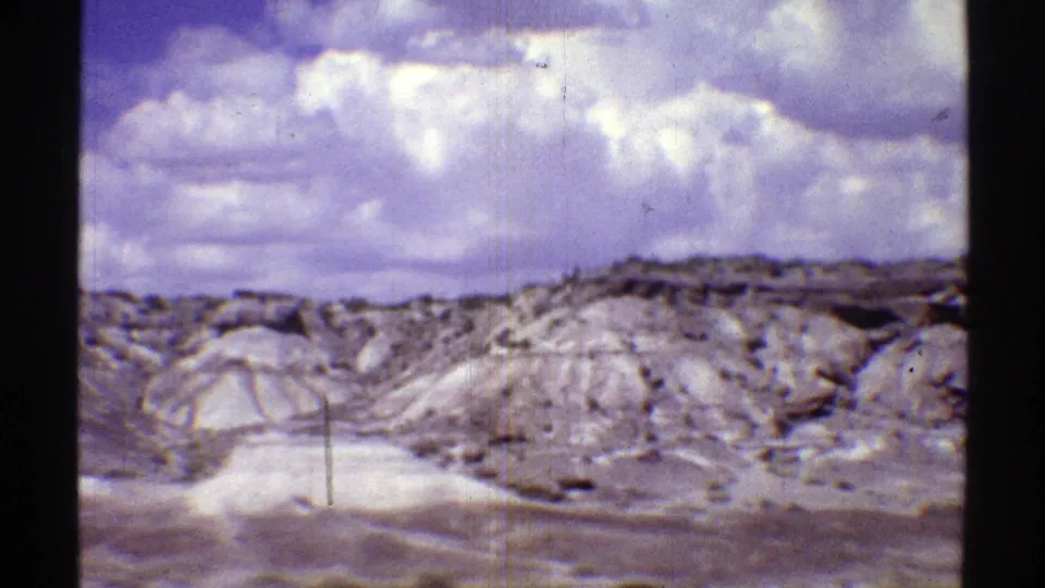 1969 Arizona home-movie frame showing pale rocky desert formations and low hills beneath large cloud cover.