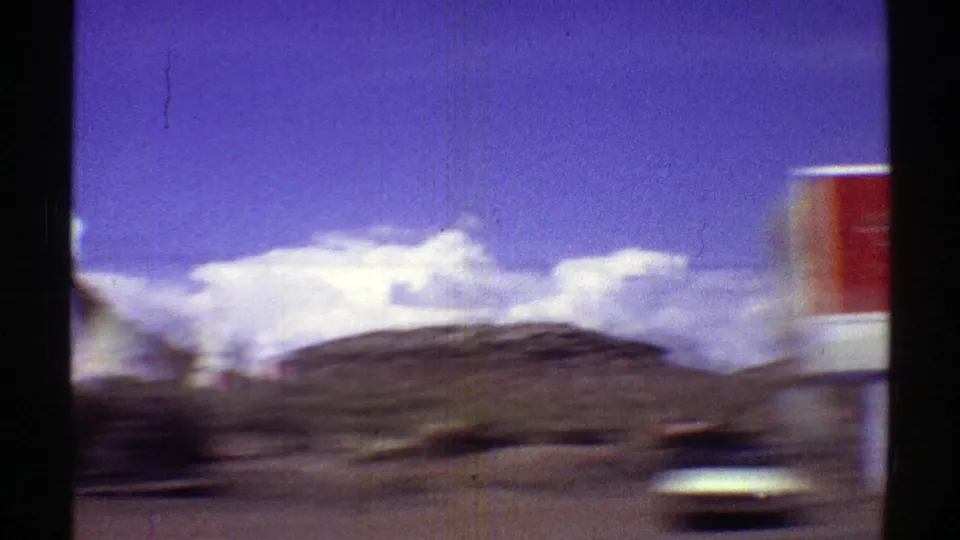A blurred moving-vehicle view passes desert roadside terrain with clouds and part of the red sign at frame edge.