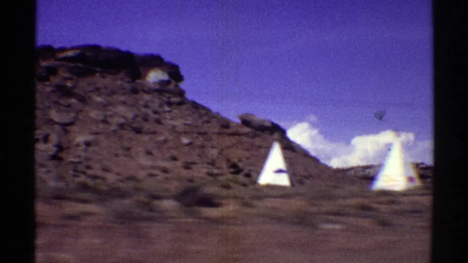 1969 Arizona home-movie frame showing white roadside teepees with red markings near a rocky desert hillside under blue sky.