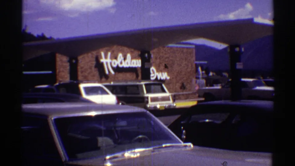 1969 home-movie frame of a Holiday Inn exterior with visible signage and several parked American cars in the lot.
