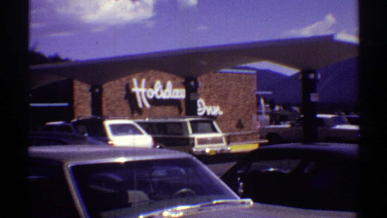 1969 Arizona 8mm frame showing a Holiday Inn facade, parked American cars, and distant mountains under a bright sky.
