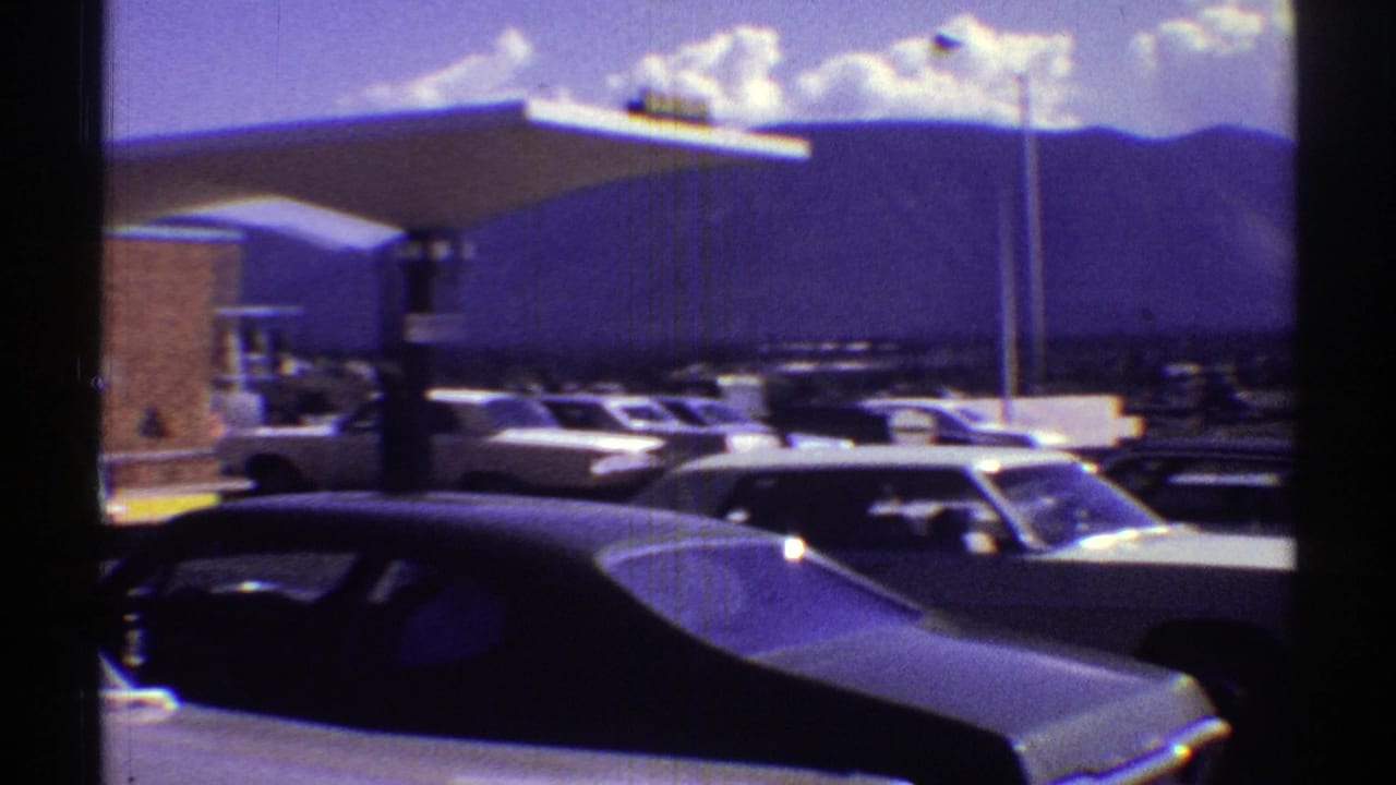 1969 Arizona frame of parked classic cars near a broad Holiday Inn canopy, with mountains and clouds beyond.