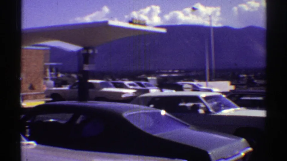 1969 Arizona frame of parked classic cars near a broad Holiday Inn canopy, with mountains and clouds beyond.