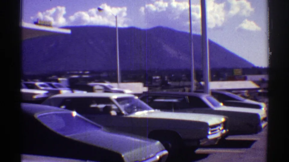 1969 Arizona 8mm view across a motel parking lot, showing classic cars, light poles, and a mountain backdrop.
