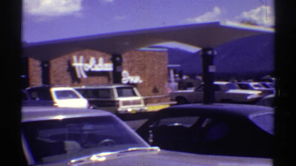 1969 Arizona frame returning to the Holiday Inn sign, brick facade, parked cars, and distant mountains.