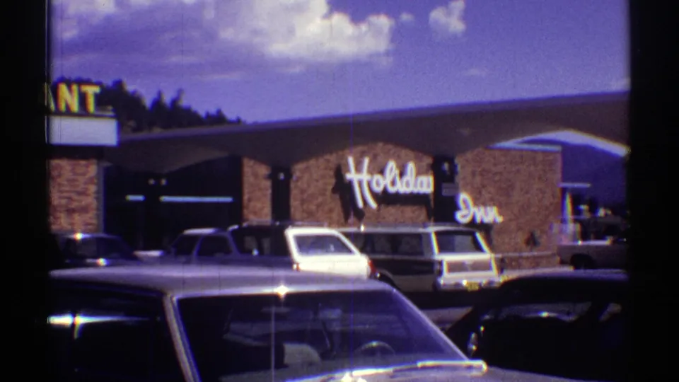 Arizona 1969 8mm frame showing the Holiday Inn sign, partial restaurant lettering, brick exterior, and parked station wagons and sedans under a clear daytime sky.
