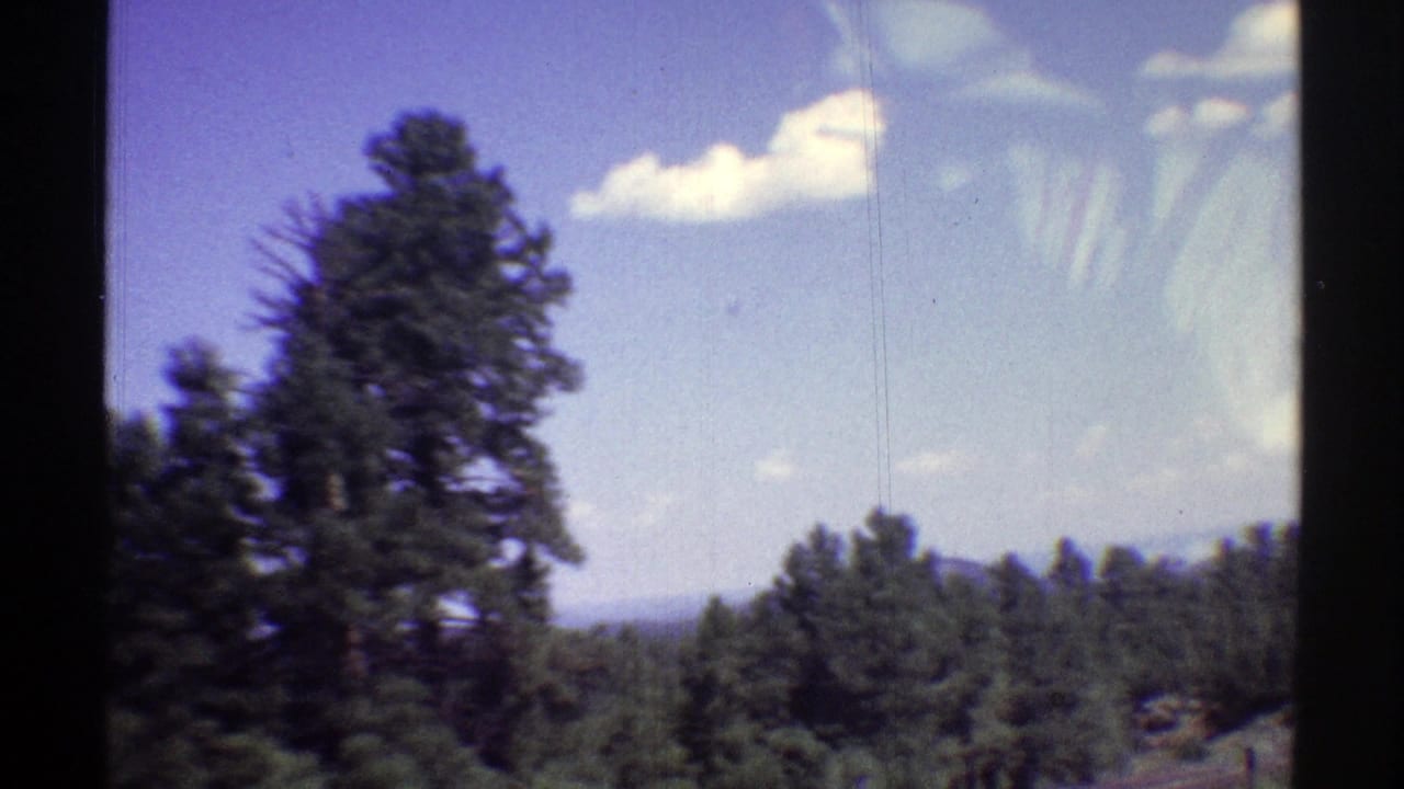 1969 Arizona home-movie frame with pine trees, distant landscape, blue sky, and small white clouds.