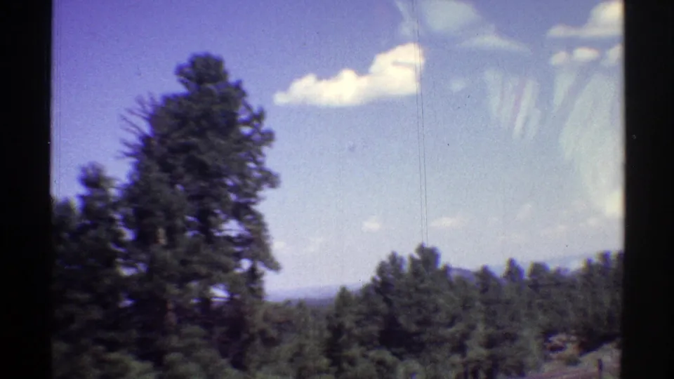 1969 Arizona home-movie frame with pine trees, distant landscape, blue sky, and small white clouds.