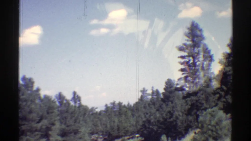 1969 Arizona 8mm frame showing roadside evergreens, a tall pine, pale blue sky, and drifting clouds.
