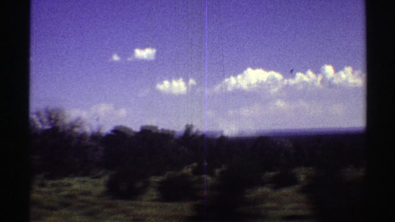 1969 Arizona travel frame with blurred roadside brush and tree shapes below a bright blue sky and horizontal cloud bank.