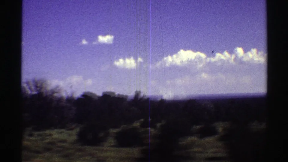 1969 Arizona travel frame with blurred roadside brush and tree shapes below a bright blue sky and horizontal cloud bank.