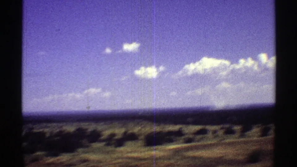 1969 Arizona 8mm frame of low scrub and green-brown field beneath scattered clouds, with visible film scratches.