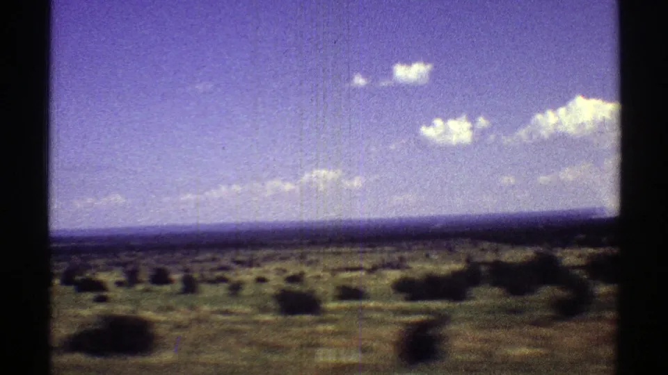 1969 Arizona landscape frame showing scattered bushes, open field, distant horizon, and bright clouds on 8mm film.