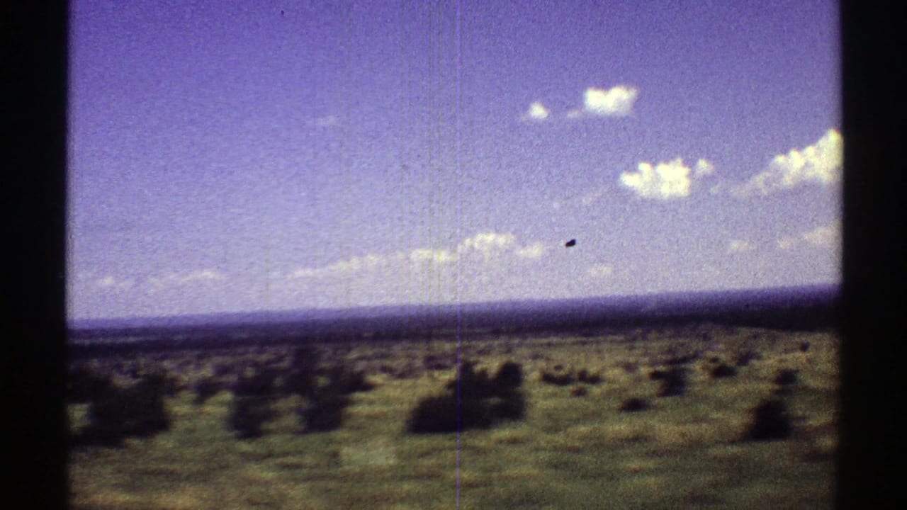 Open scrubland under a blue-purple sky with small white clouds and dark film gate edges.
