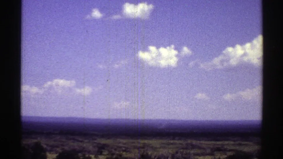 1969 Arizona landscape frame with sparse desert vegetation, a long horizon, and bright clouds in a blue-purple sky.