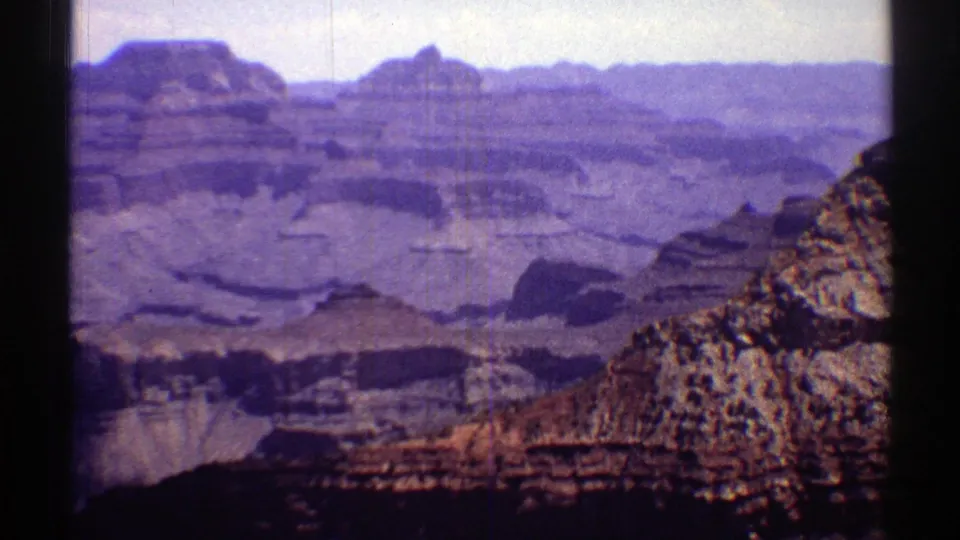 1969 Arizona 8mm frame showing layered canyon formations, distant mesas, and a rocky foreground slope under a pale sky.
