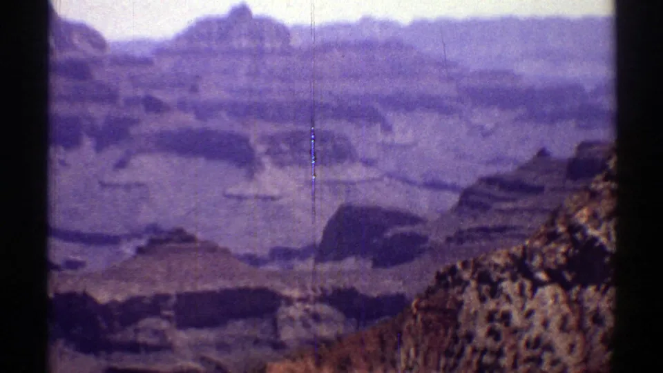 1969 8mm Arizona canyon view with distant layered cliffs, a rocky foreground ridge, soft focus, and dark frame borders.
