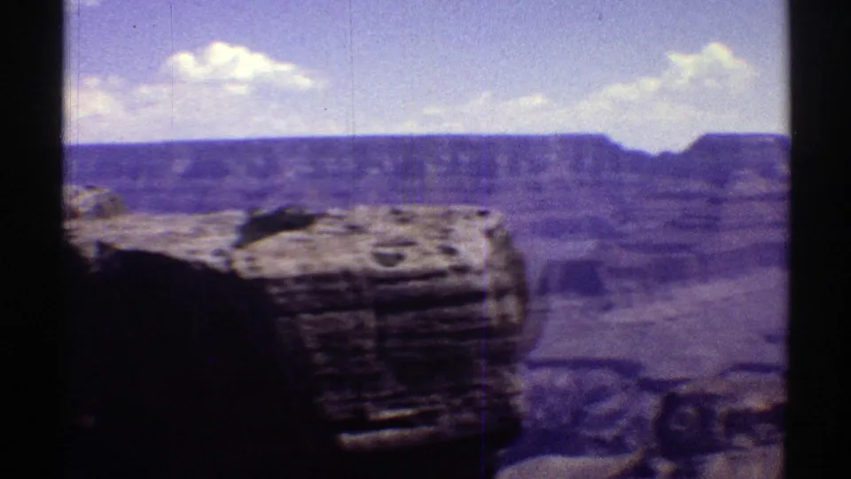 1969 Arizona travel footage frame with a foreground rock ledge, distant canyon rim, and bright clouds softened by 8mm grain.