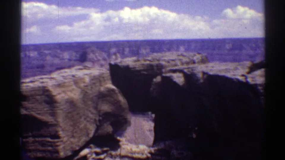 Arizona canyon overlook on 8mm film, with rough foreground rock, distant layered cliffs, and a cloud band across the sky.