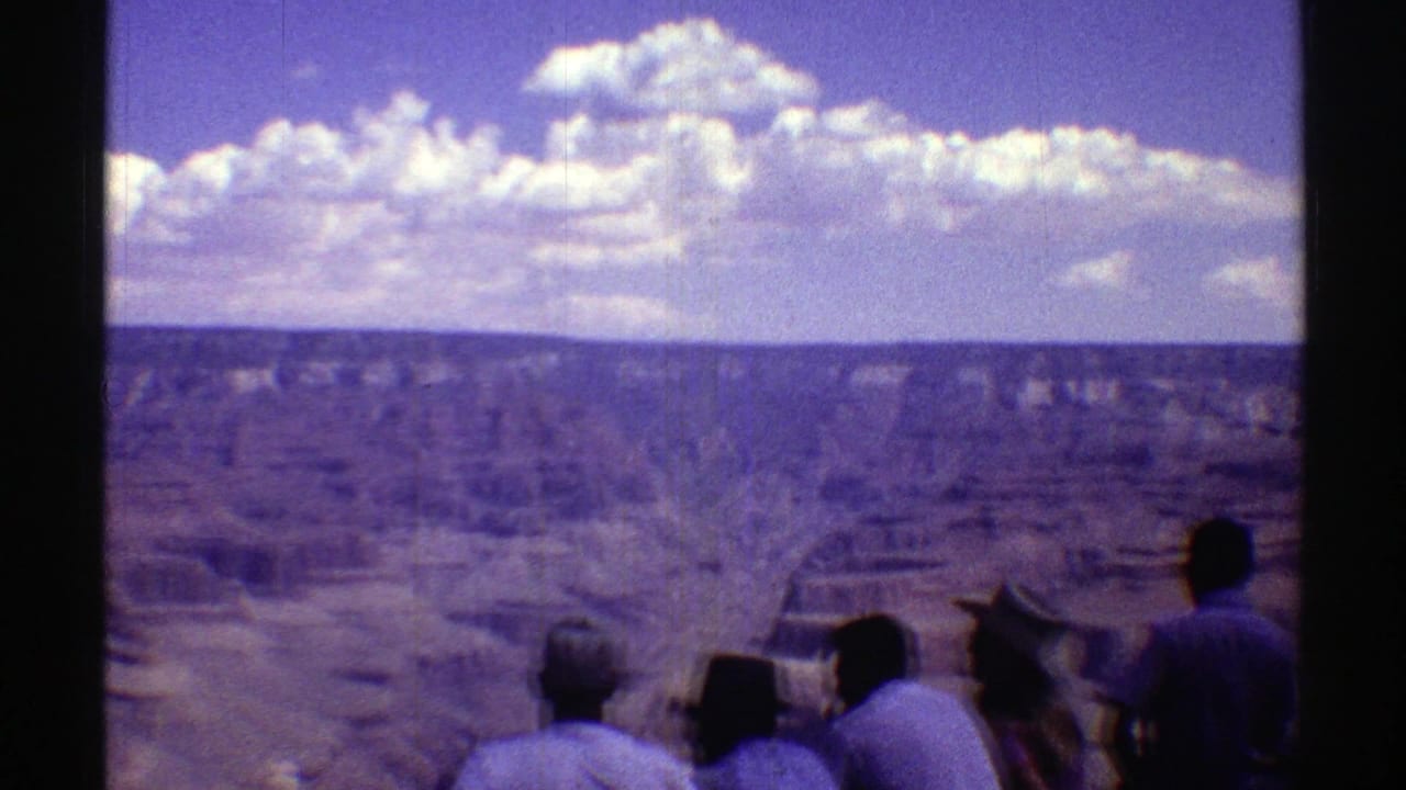 People at an overlook face a wide canyon with clouds above the horizon.