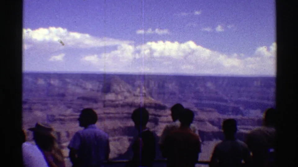 Late-1960s Arizona home-movie frame of silhouetted visitors at a canyon viewpoint under a bright cloud line.