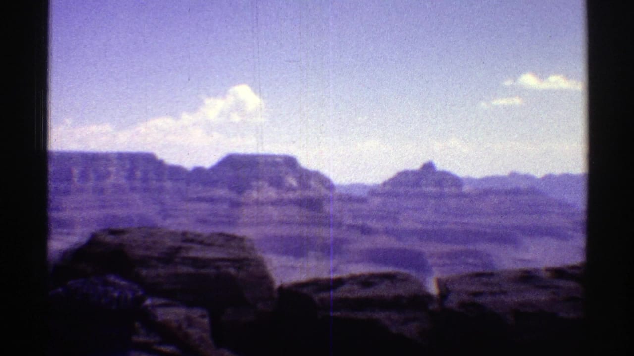 Grand Canyon view with dark foreground rocks, layered cliffs, blue sky, and scattered clouds.