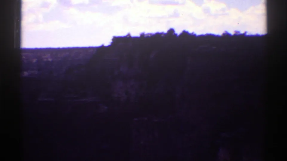 1969 Arizona 8mm frame showing a dark canyon rim, steep cliff face, bright clouded sky, and deep shadow across the terrain.