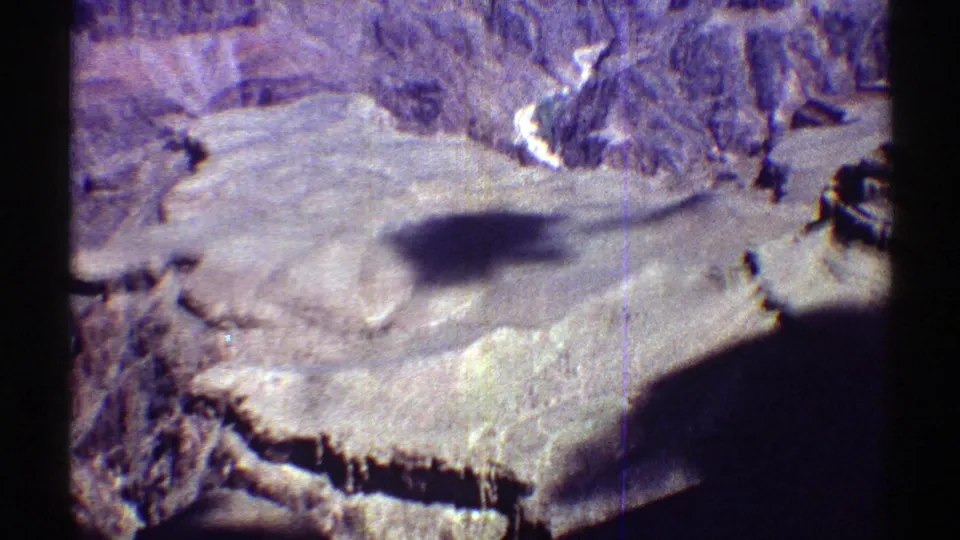 1969 Arizona 8mm aerial frame showing pale canyon plateau, eroded ravines, vertical rock walls, and a dark obstruction at the lower edge.