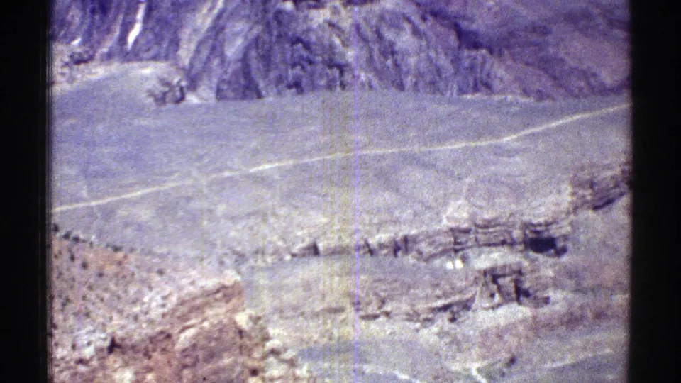 Aerial 8mm frame over Arizona highlands, with a pale road, rocky slopes, scrub vegetation, and distant rugged hills.