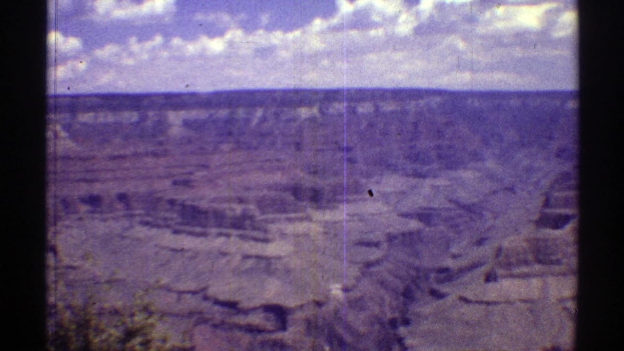 Wide canyon landscape with layered rock, distant mesa, blue sky, and scattered clouds.
