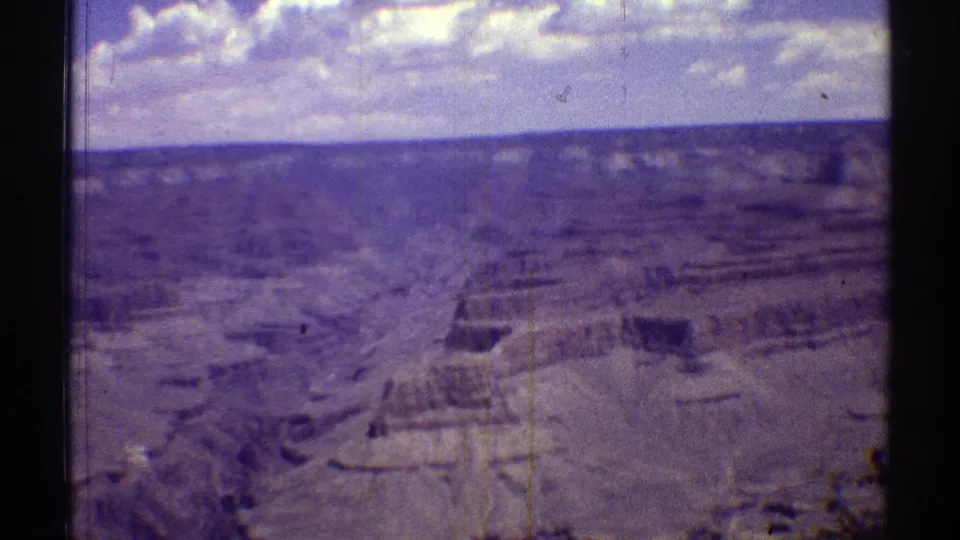 1969 Arizona 8mm frame showing a soft-focus view across layered canyon cliffs, pale terraces, and a bright clouded sky.
