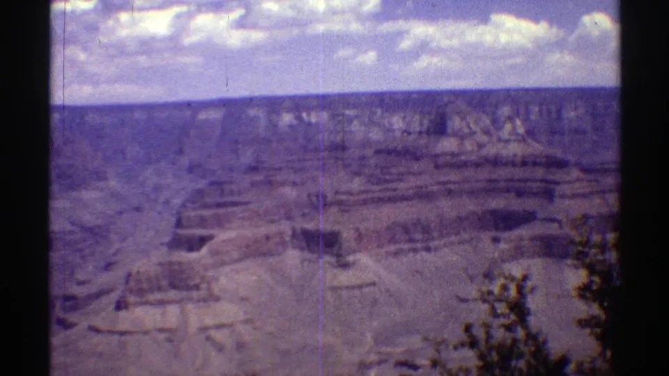 1969 Arizona home-movie frame with canyon terraces, a distant plateau, clouded blue sky, and sparse trees at the edge.