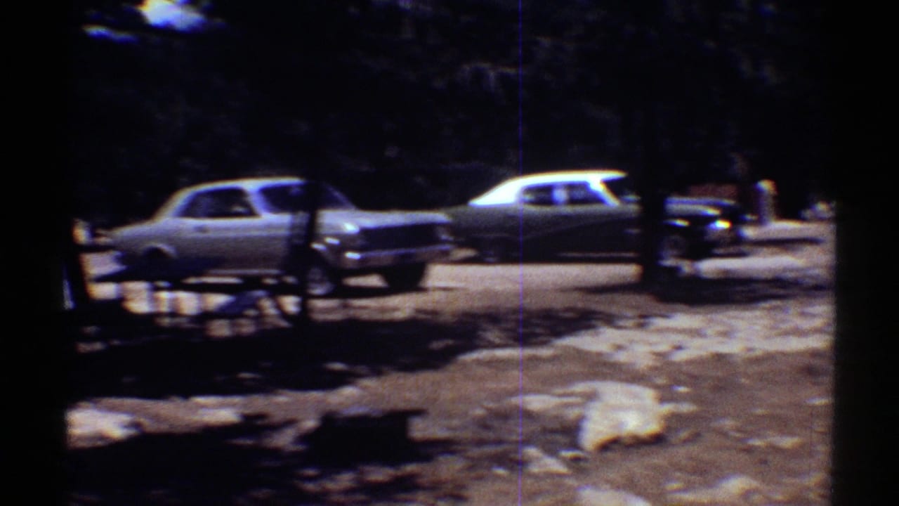 Shaded parking area with vintage cars, trees, benches or railings, and patchy ground.