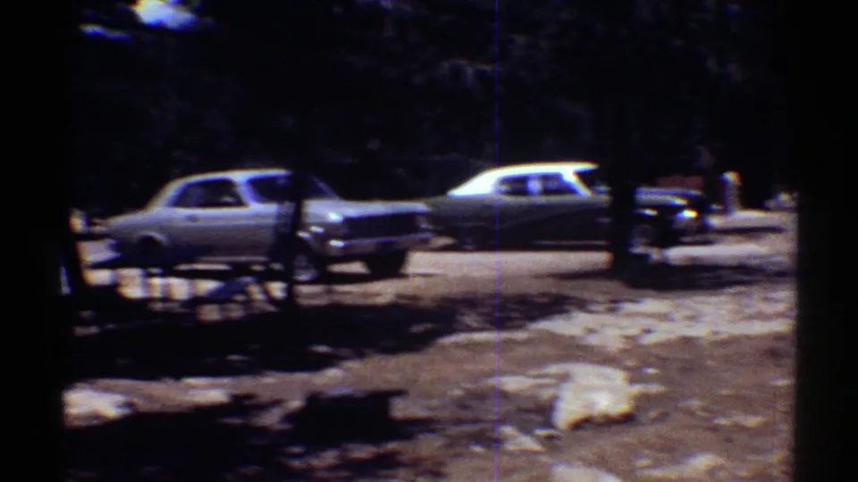 1969 Arizona home-movie frame showing parked mid-century cars in a wooded visitor lot with dark tree shadows.
