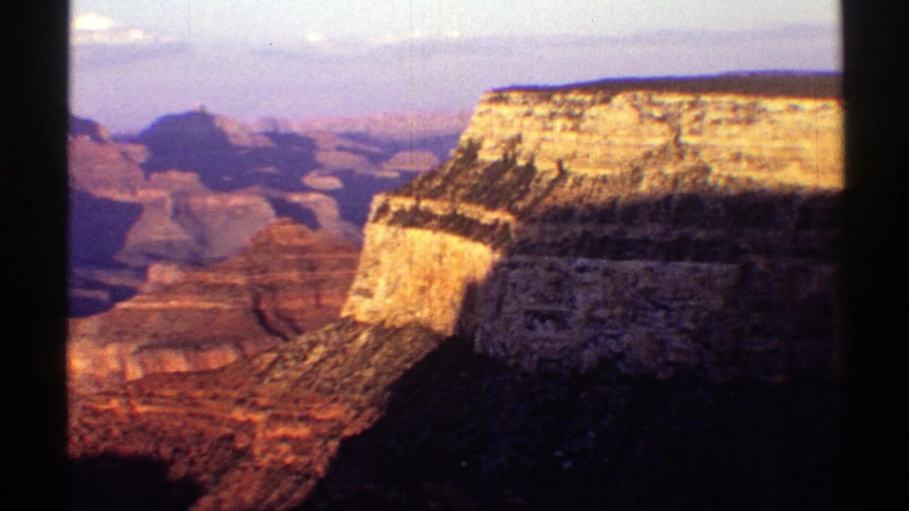 1969 8mm Arizona footage showing a Grand Canyon overlook with sunlit rock layers, dark ravines, pale sky, and visible scan edges.