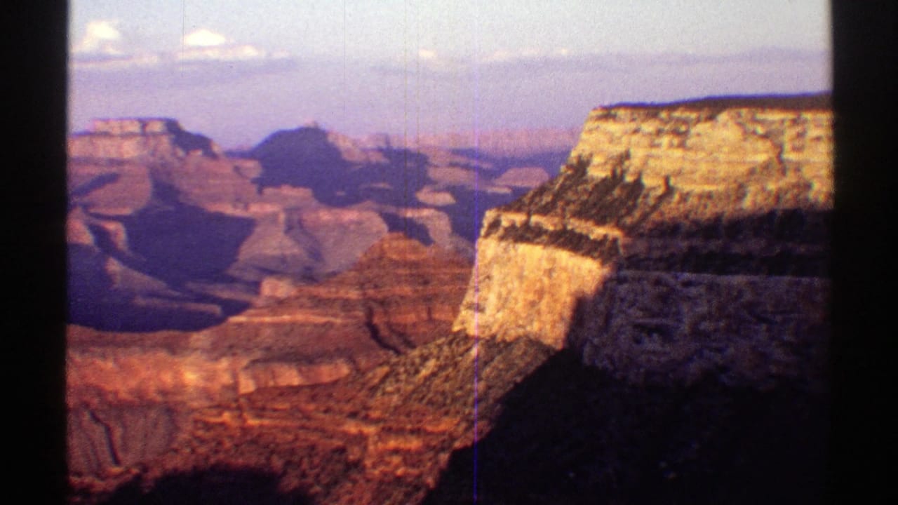 1969 8mm Arizona view of the Grand Canyon, with a bright cliff face at right, distant mesas, red rock layers, and light film scratches.