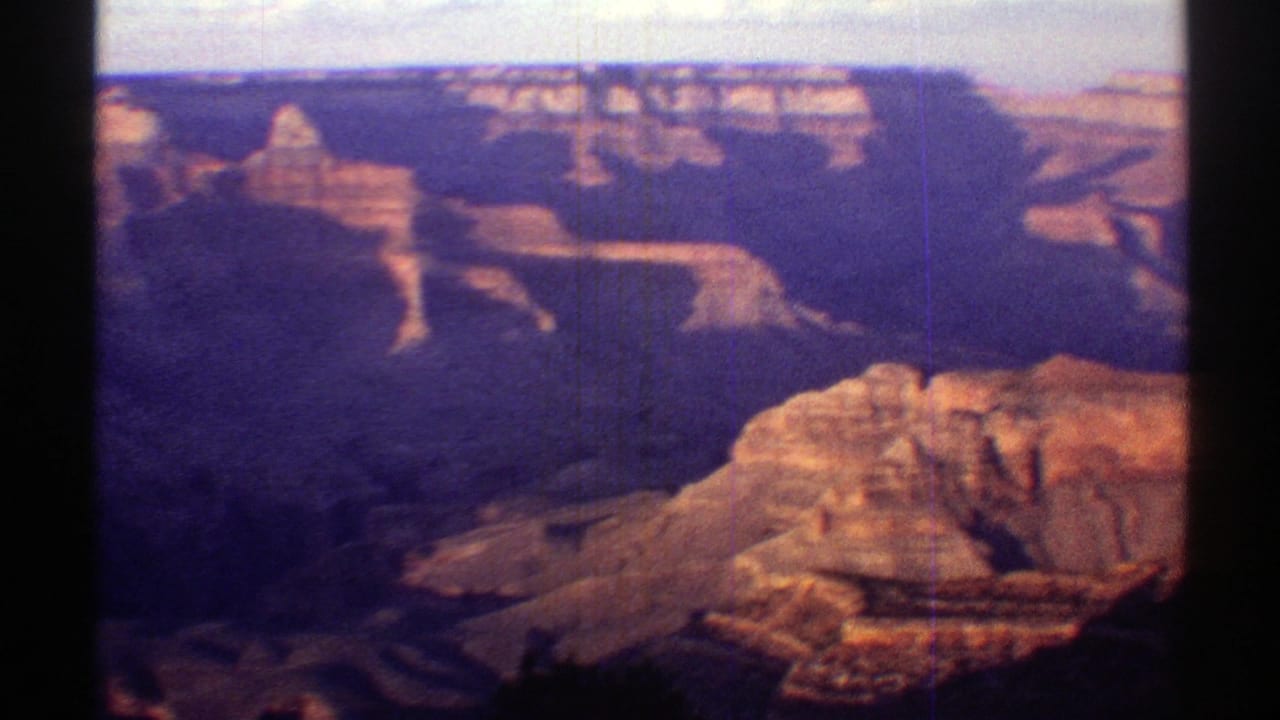 Vintage 8mm Grand Canyon panorama showing broad shadowed canyon depth, orange rock ledges, and a pale morning sky.