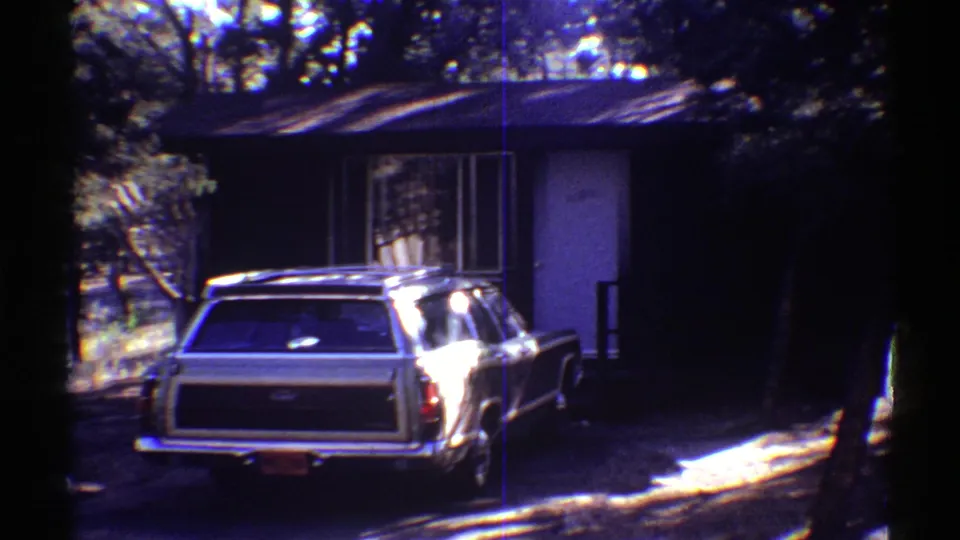 Rear of a 1960s station wagon near a cabin door and window, filmed in shaded woodland light.